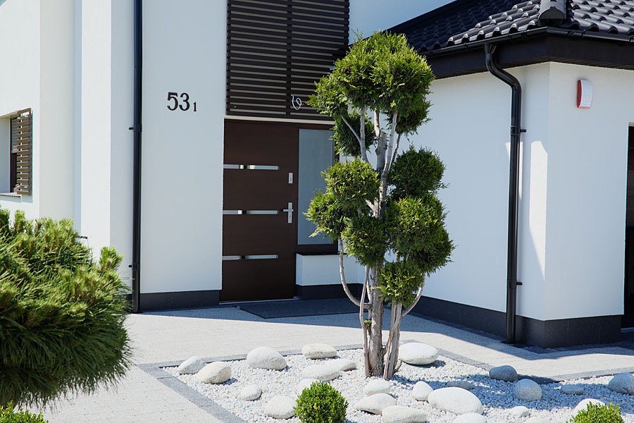 Nowoczesne wejście do domu z brązowymi drzwiami z poziomymi przeszkleniami, numerem domu 53 i ozdobnym drzewkiem w białych kamieniach.