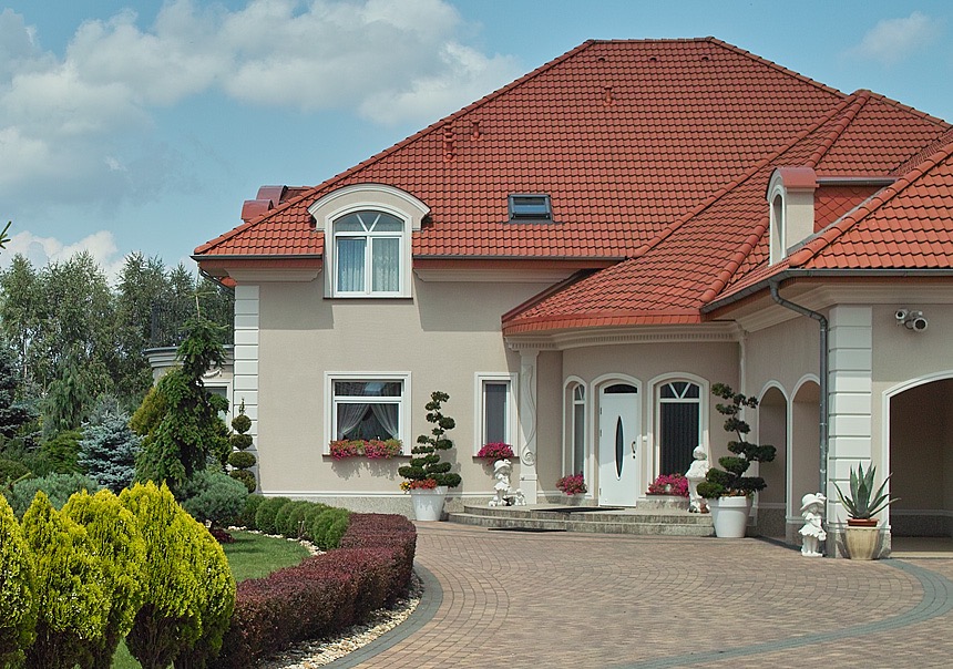 Elegancki dom jednorodzinny z białymi ramami okiennymi i wejściowymi, czerwonym dachem, zadbanym ogrodem z krzewami i drzewami ozdobnymi oraz brukowanym podjazdem.