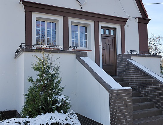 Wejście do domu z brązową, drewnianą konstrukcją, schodami z cegły i małym drzewkiem iglastym w śniegu.