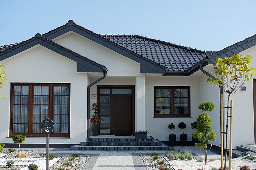 Elewacja nowoczesnego domu jednorodzinnego z ciemnymi oknami i wejściem, ciemna dachówka, białe ściany, zagospodarowany ogród z kamykami i niskimi roślinami, skrzynka pocztowa z napisem 'Listy'.