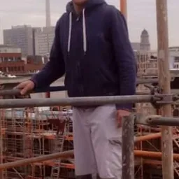 Pracownik budowlany w ubraniu roboczym stoi na rusztowaniu z widokiem na panoramę Berlina z wieżą telewizyjną w tle.