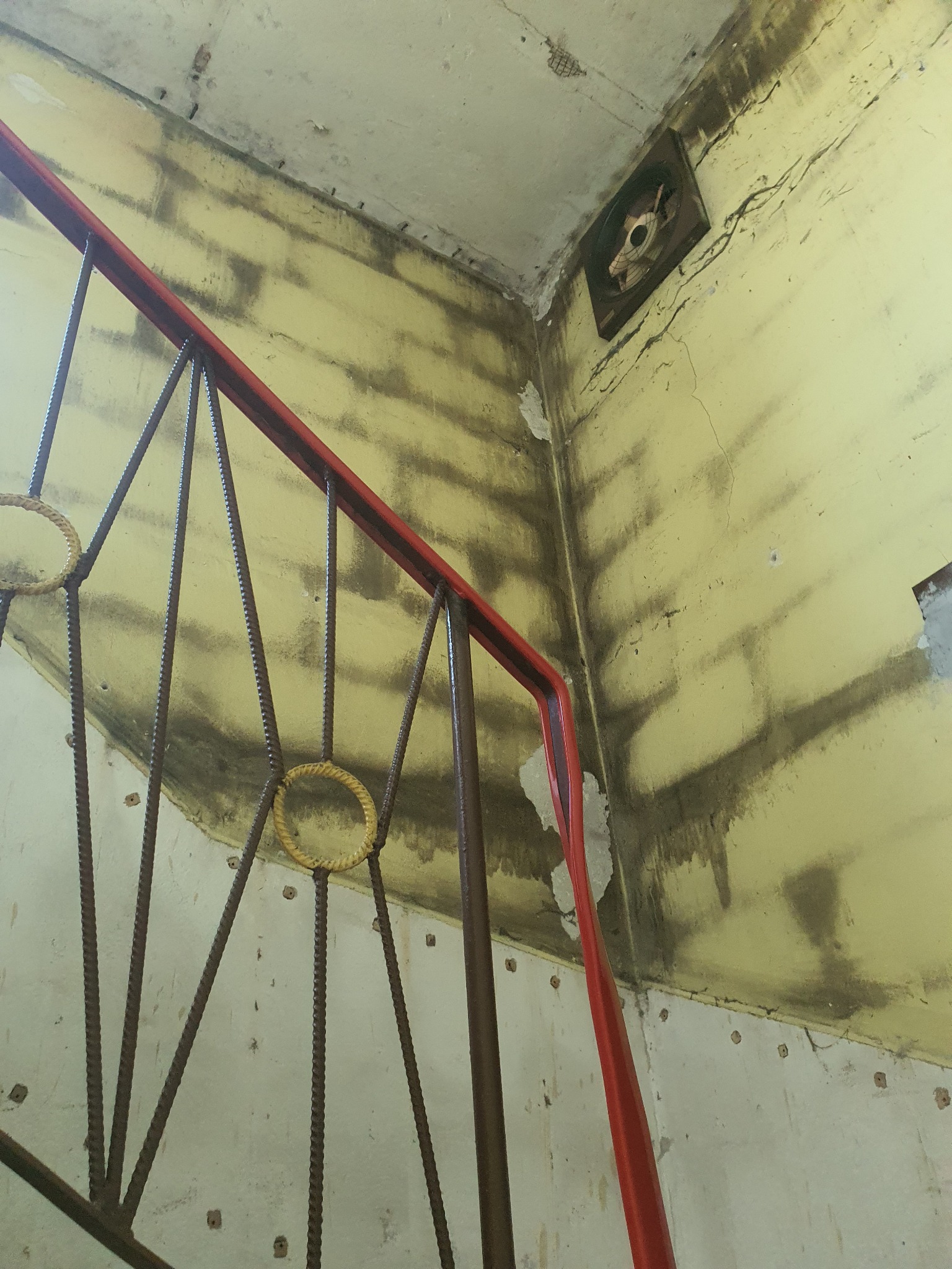 Klatka schodowa w trakcie remontu, widoczne stare, żółte ściany z zaciekami i uszkodzeniami, metalowa balustrada z czerwonymi elementami i ozdobnymi, okrągłymi detalami z grubego sznura, na ścianie...