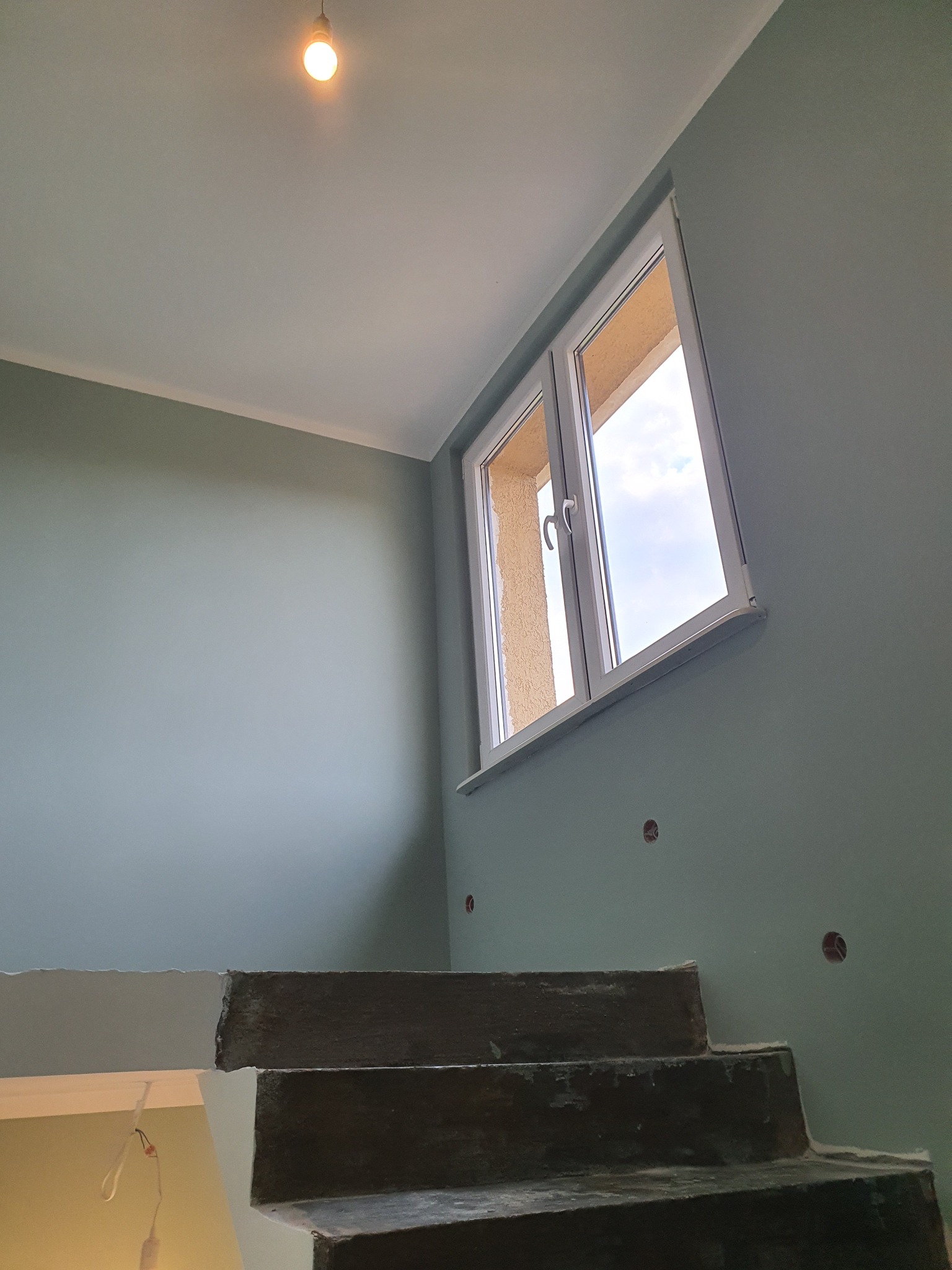 Świeżo pomalowane wnętrze w odcieniach szarości z surowymi, betonowymi schodami prowadzącymi w górę, widoczne gniazdka elektryczne bez osprzętu i okno z widokiem na niebo.