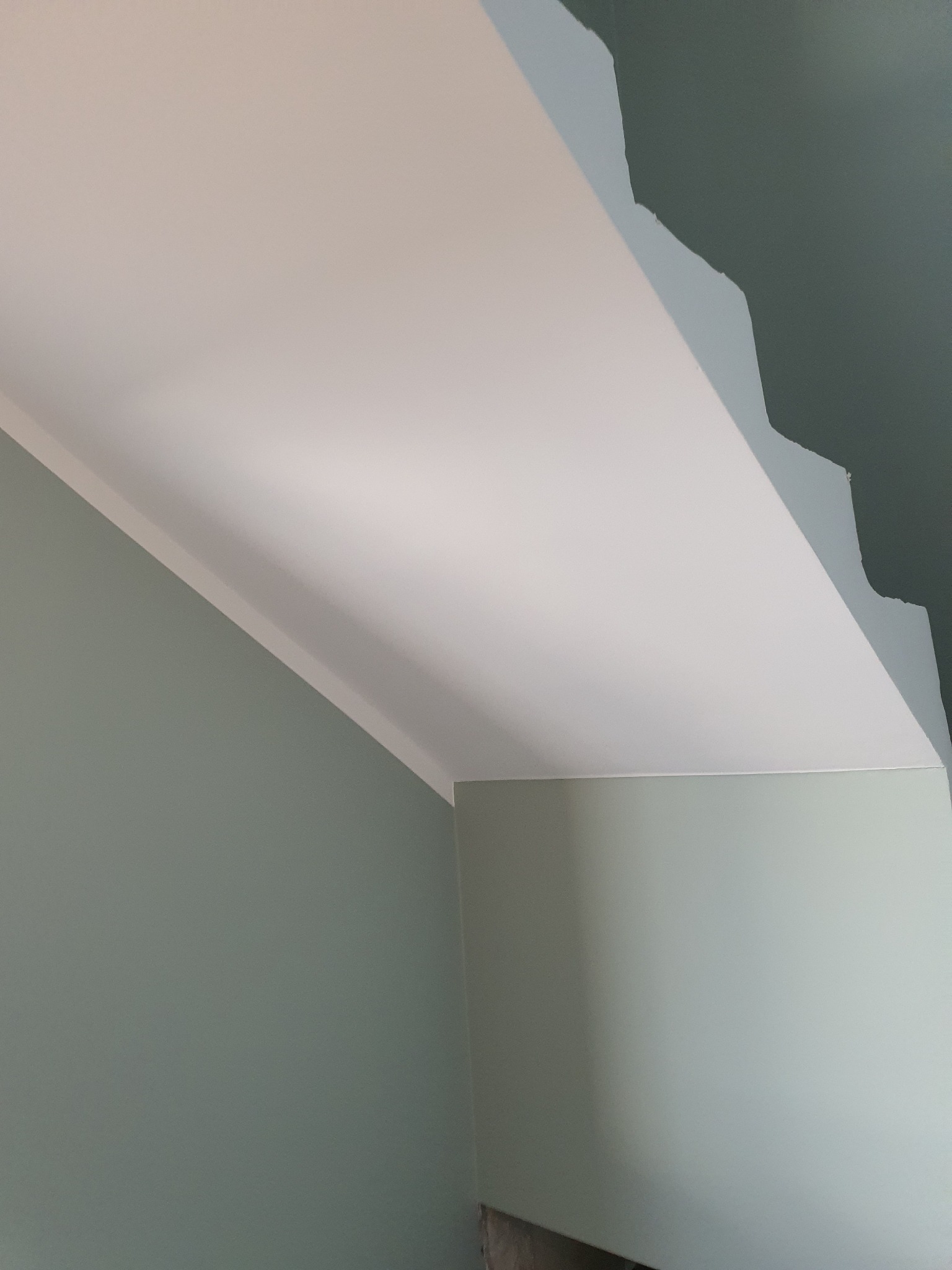 Świeżo pomalowany narożnik pomieszczenia z szarozielonymi ścianami i białym sufitem, widoczne nierówności na suficie przed ostatecznym wykończeniem.