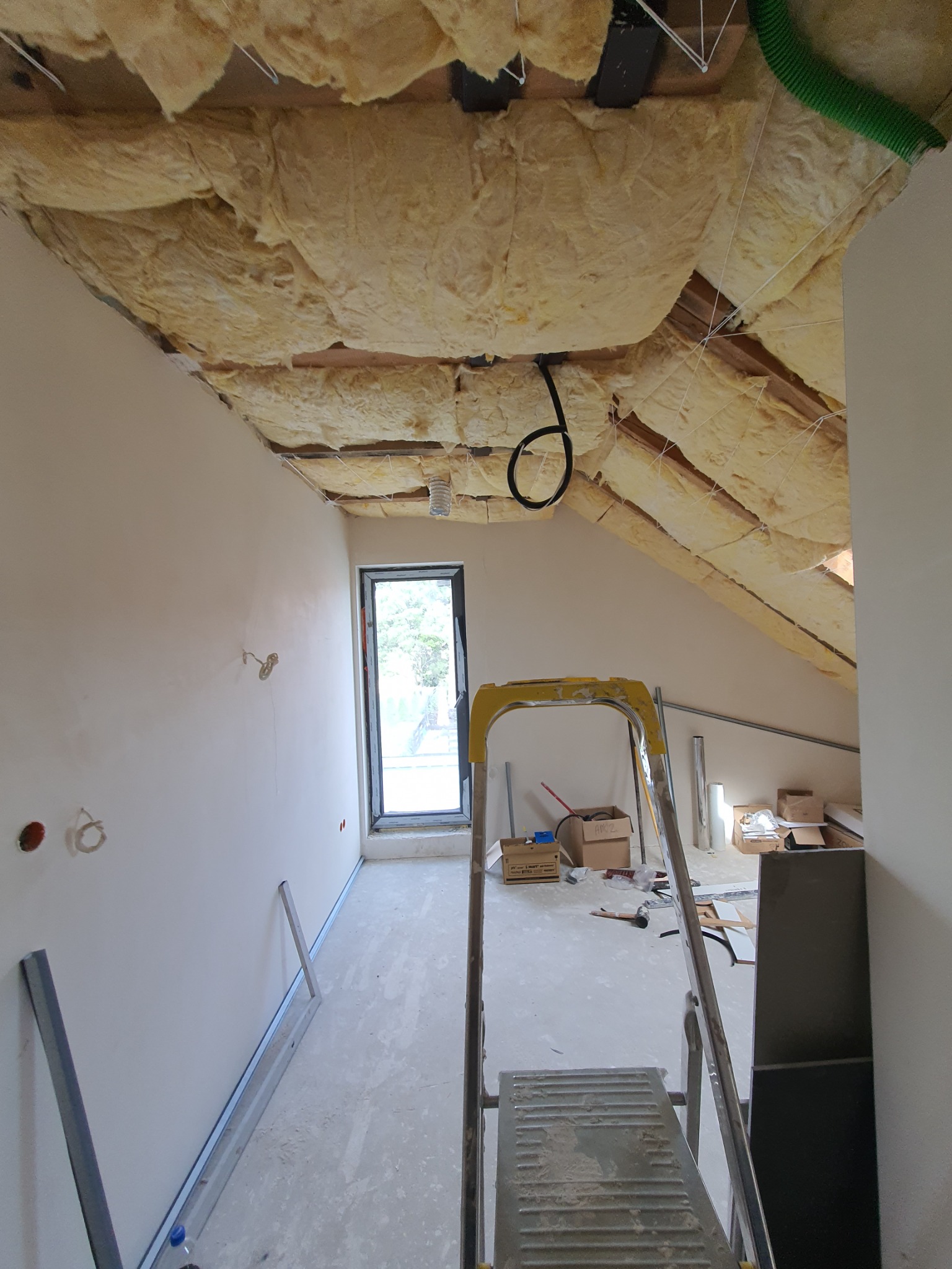 Poddasze w trakcie prac wykończeniowych: ocieplenie wełną mineralną poddasza zabezpieczone sznurkami, ściany przygotowane pod malowanie, widoczne profile metalowe, drabina i okno dachowe.