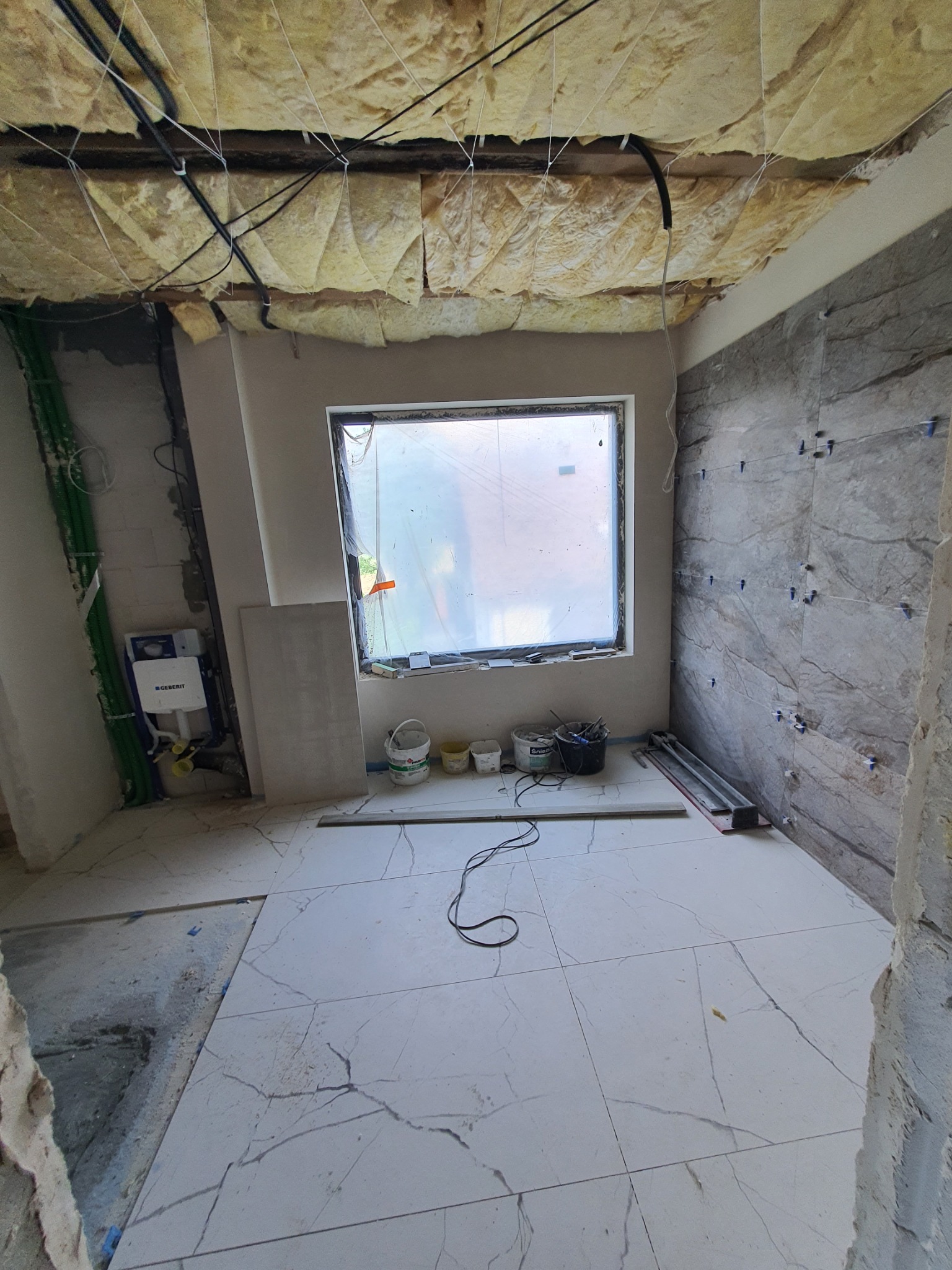 Wnętrze łazienki w trakcie remontu: położone białe płytki podłogowe z szarymi wzorami, ściana częściowo wyłożona szarą płytką imitującą kamień z niebieskimi krzyżykami dystansowymi, odsłonięta...