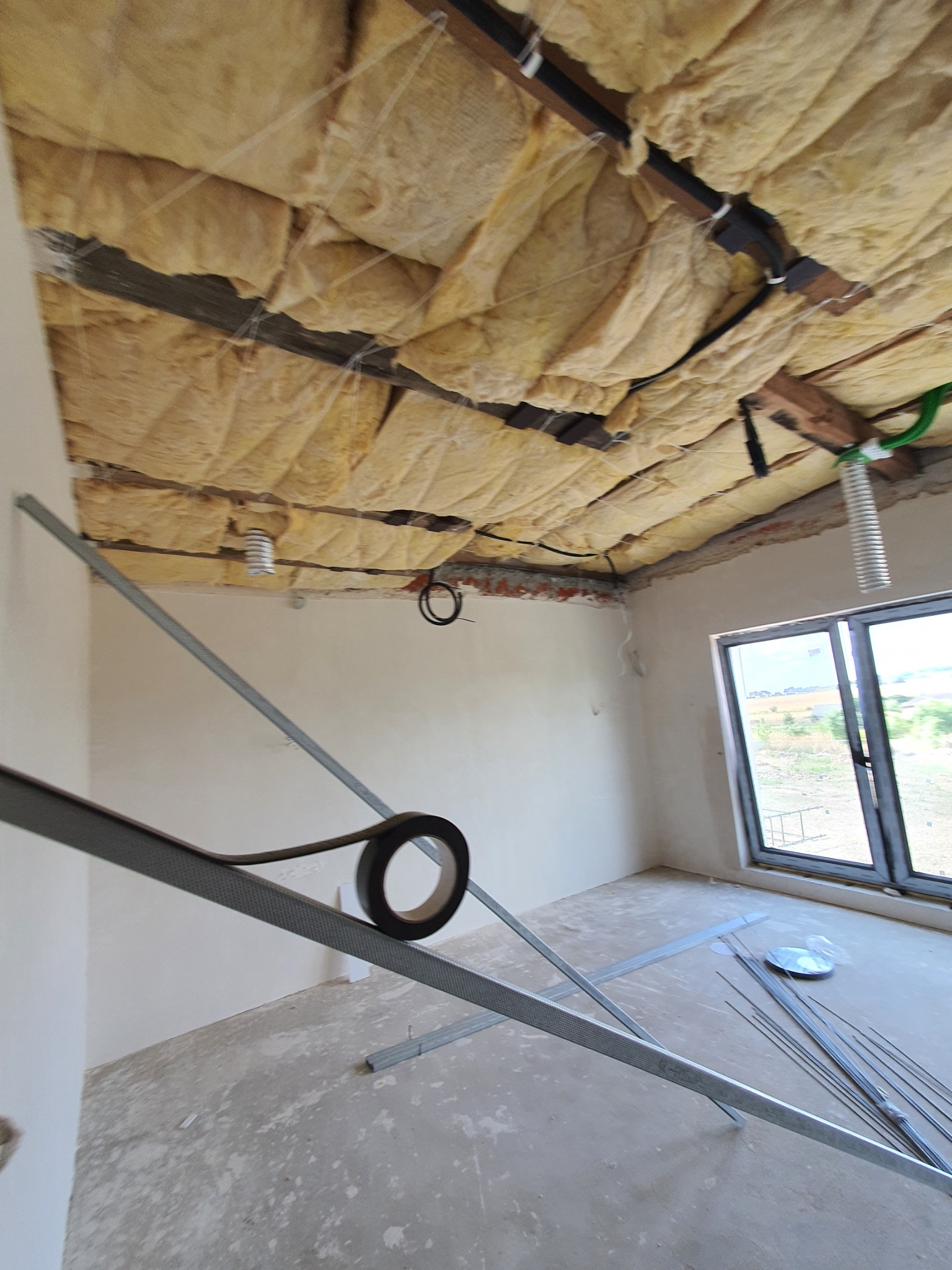 Montaż izolacji stropu wełną mineralną, widoczne profile stalowe, taśma uszczelniająca, okno i surowy beton na podłodze.