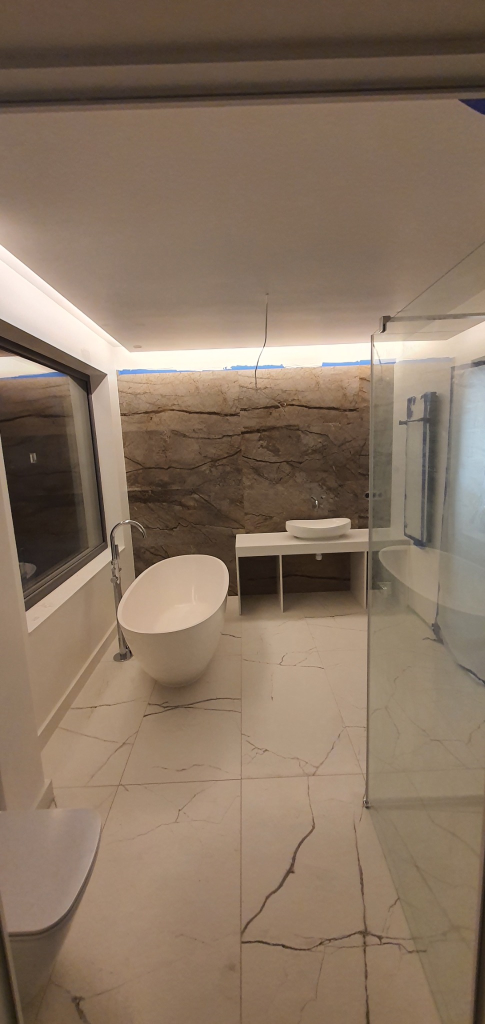 Nowoczesna łazienka z wolnostojącą wanną, umywalką nablatową na białej szafce i szklaną kabiną prysznicową; ściana wykończona płytkami imitującymi marmur, widoczne niedokończone prace przy...