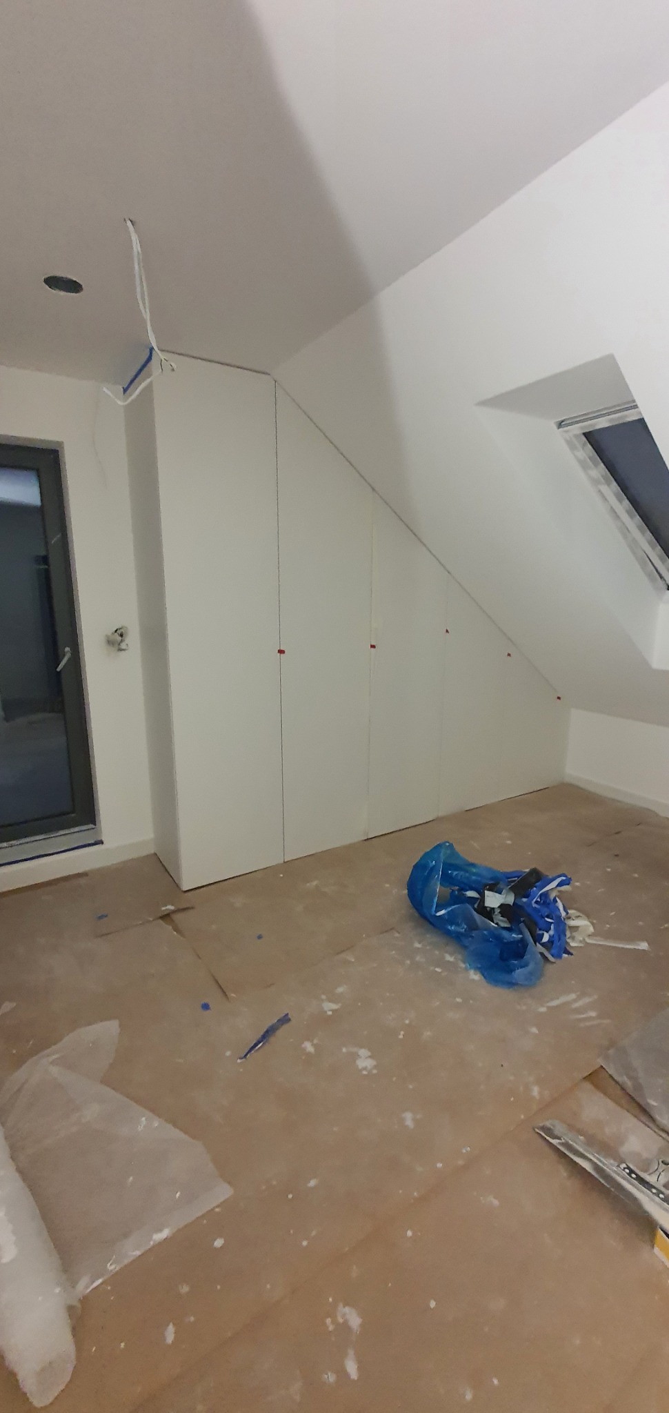 Poddasze w trakcie prac wykończeniowych: zabudowa szaf wnękowych pod skosem, zabezpieczona podłoga kartonem, widoczne narzędzia i odpady w niebieskim worku.