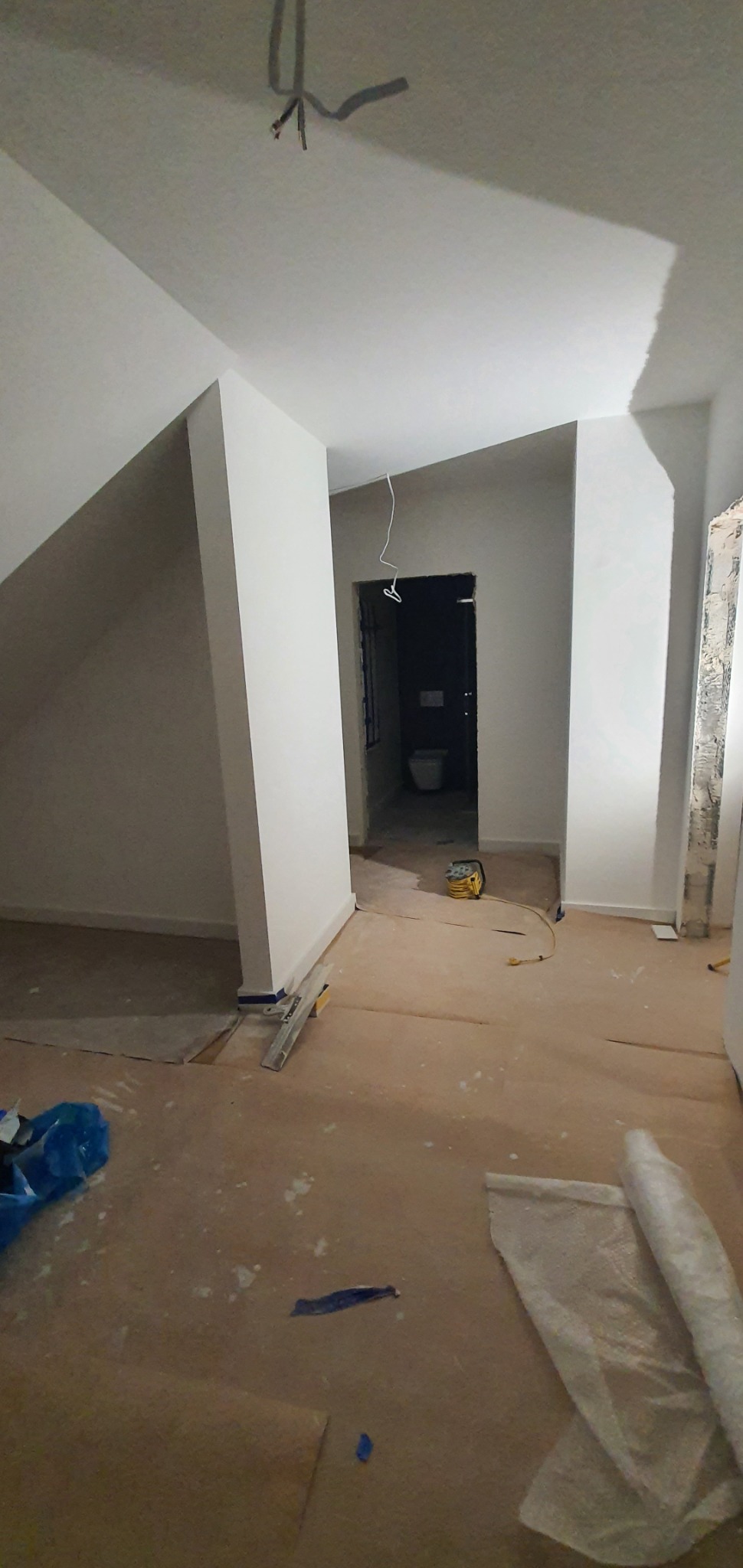 Wnętrze w trakcie prac wykończeniowych: świeżo pomalowane ściany na biało, zabezpieczona podłoga, widoczne narzędzia malarskie i przewody elektryczne wystające z sufitu, zarys łazienki w tle.
