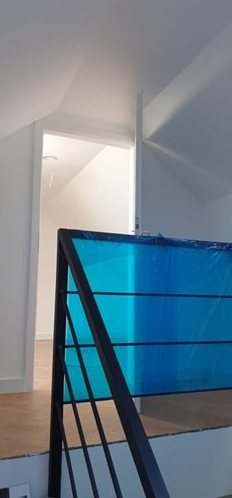 Klatka schodowa zabezpieczona niebieską folią ochronną podczas prac wykończeniowych, widoczny fragment czarnej, metalowej balustrady i białe ściany poddasza.