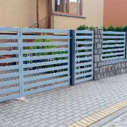 Nowoczesne ogrodzenie panelowe w kolorze szarym z poziomymi szczeblami, osadzone na kamiennej podmurówce, z widoczną bramą wjazdową i fragmentami budynków w tle.