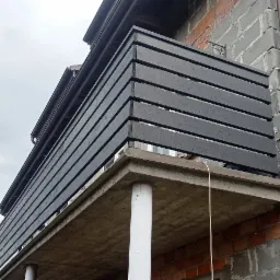 Czarny, drewniany balkon z poziomymi deskami, widoczny z dołu, na tle surowej ściany budynku z cegły i betonu.