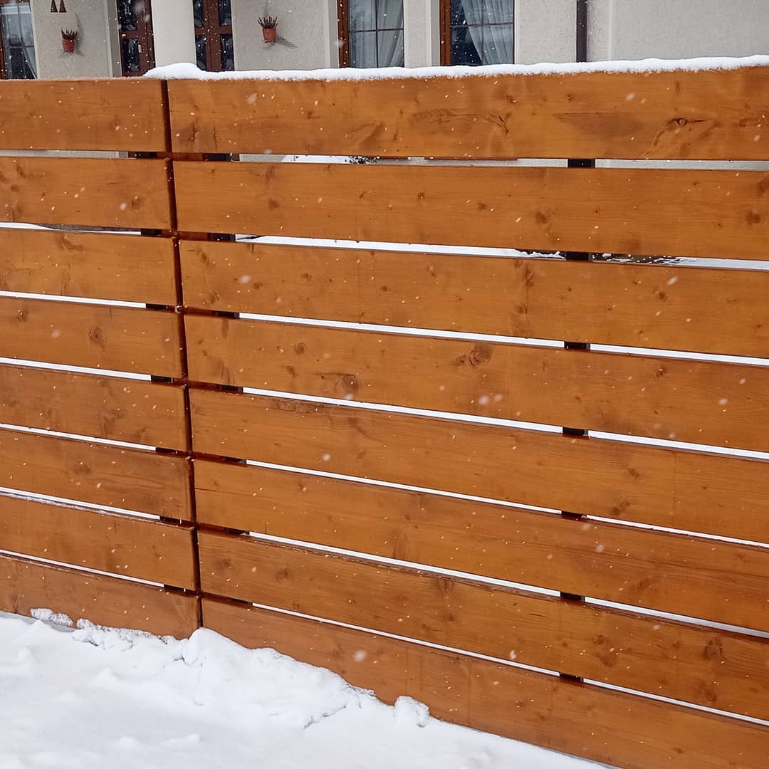 Nowoczesne ogrodzenie z poziomych desek w ciepłym odcieniu brązu, częściowo pokryte śniegiem, z widocznymi śladami opadów śniegu w powietrzu i fragmentem budynku w tle.