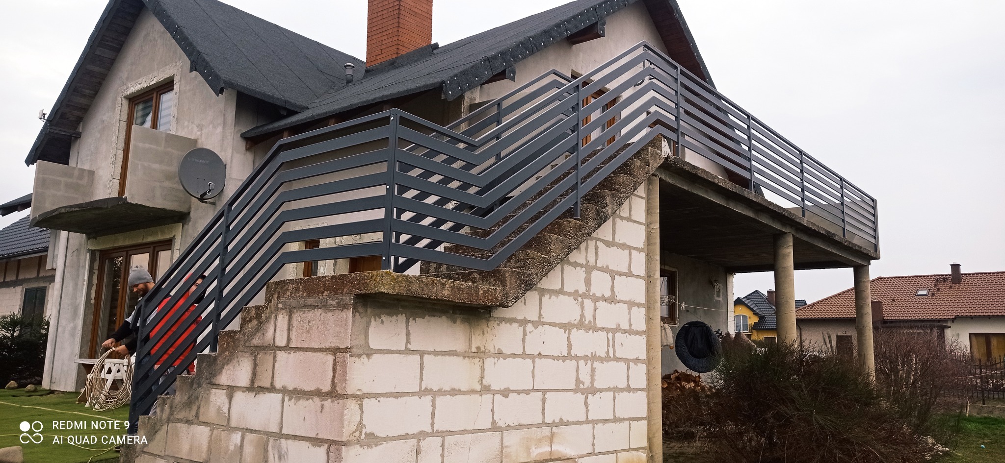 Zewnętrzne schody betonowe z nowoczesną, metalową balustradą w kolorze grafitowym, prowadzące na balkon domu w trakcie budowy, widoczne osoby pracujące przy instalacji.