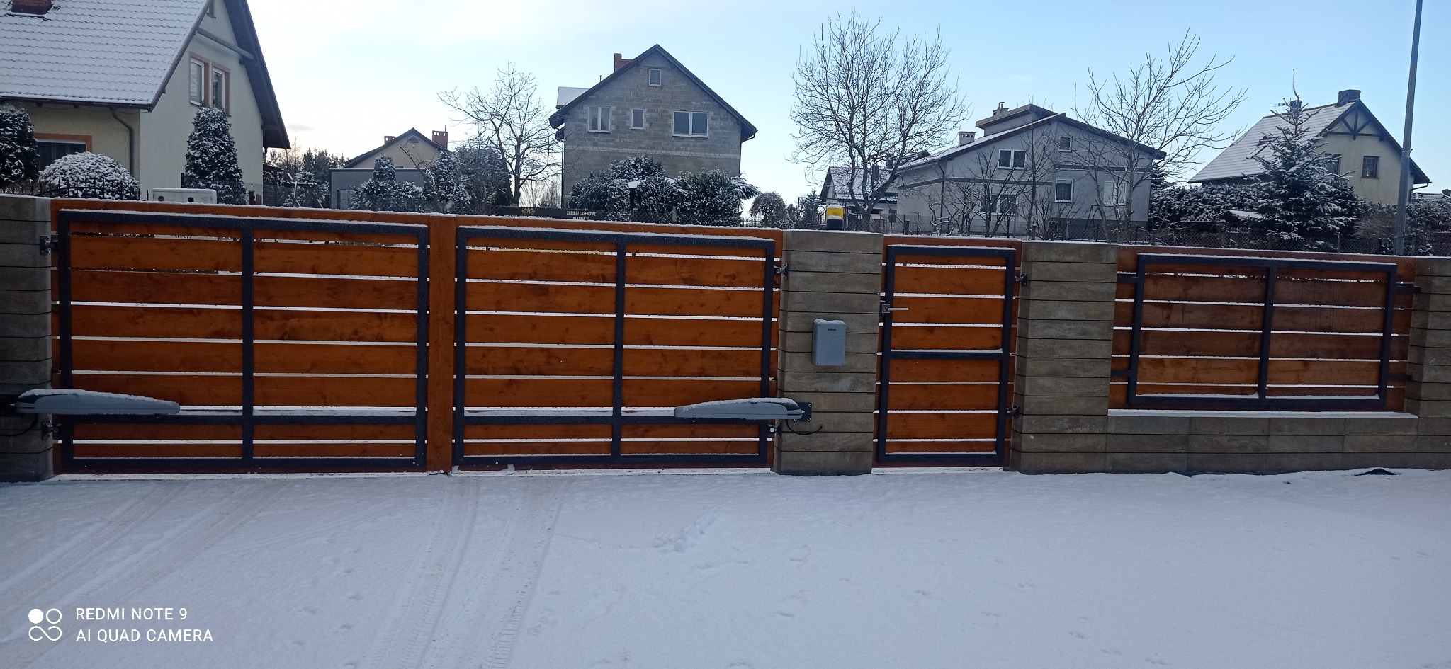 Nowoczesna brama wjazdowa z automatyką, drewniane wypełnienie, metalowa rama, słupki z szarej kostki, zimowy krajobraz z ośnieżonym podjazdem i domami w tle.