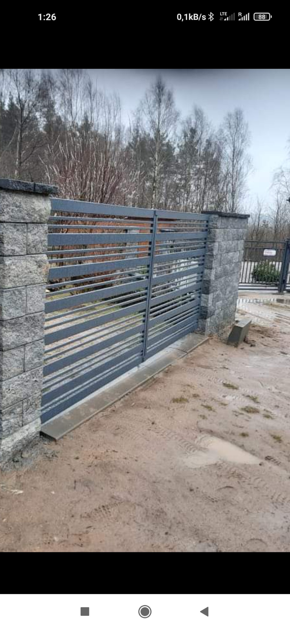 Nowoczesna, pozioma brama wjazdowa z szarych, metalowych profili, osadzona na słupkach z szarej kostki, na tle piaszczystego podłoża i drzew bez liści.