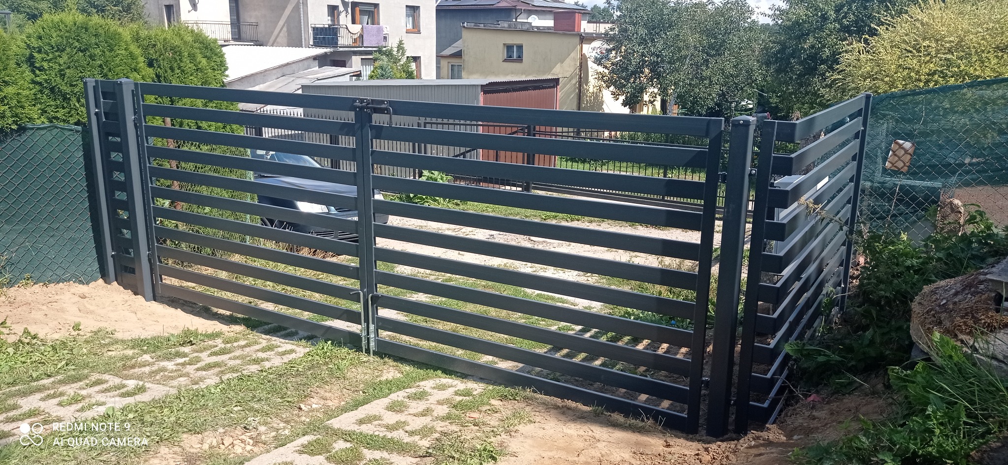 Nowoczesna, pozioma brama wjazdowa z metalowych profili pomalowanych na grafitowo, z widocznym mechanizmem zamykającym, na tle posesji z kostką brukową i zielenią.