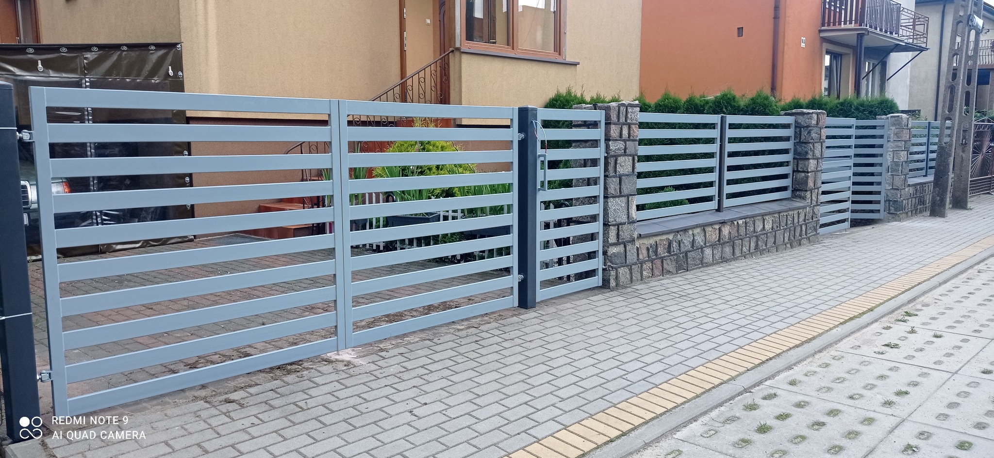 Nowoczesne ogrodzenie panelowe w kolorze szarym z poziomymi szczeblami, osadzone na kamiennej podmurówce, z widoczną bramą wjazdową i fragmentami budynków w tle.