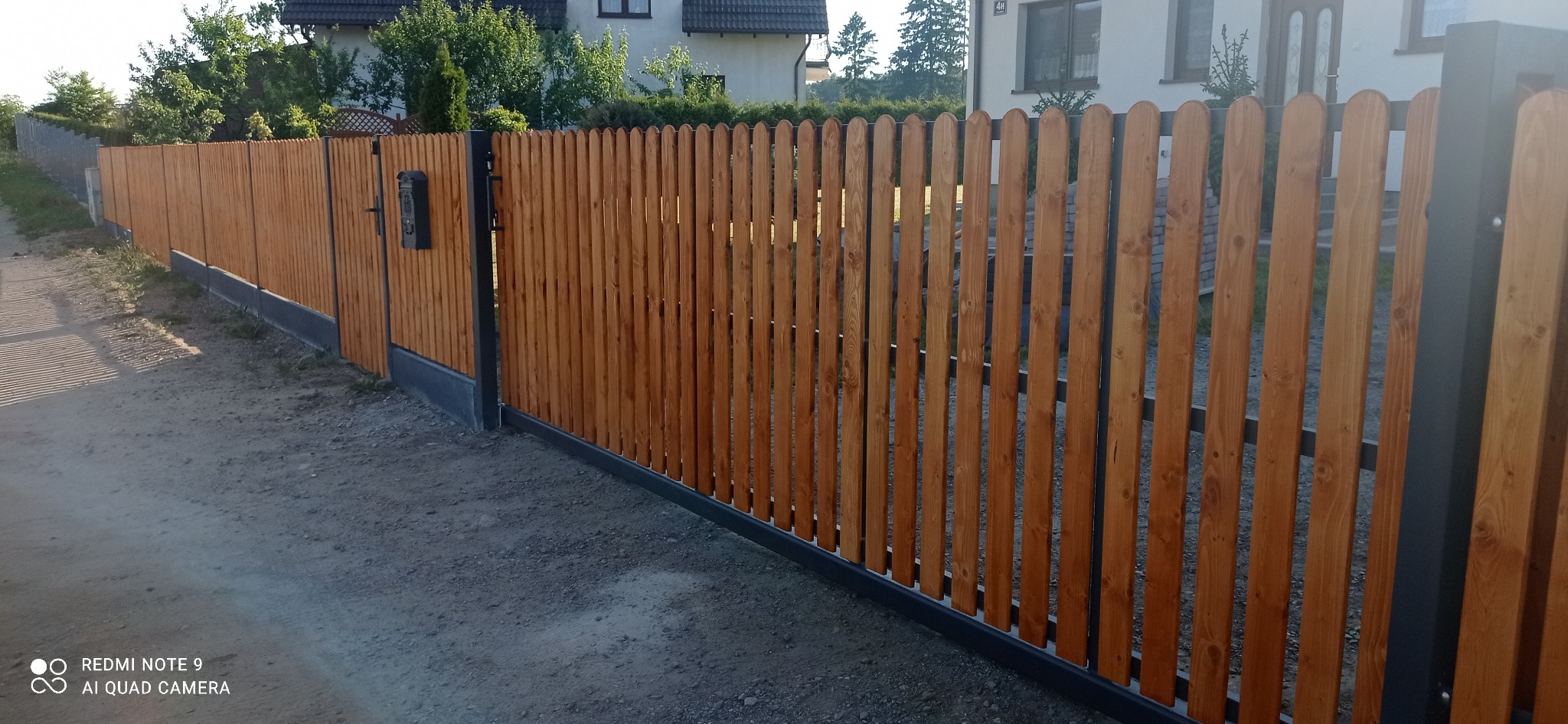 Długie, drewniane ogrodzenie palisadowe z metalowymi słupkami i podmurówką, widoczne z perspektywy drogi gruntowej, z zabudową mieszkalną w tle.