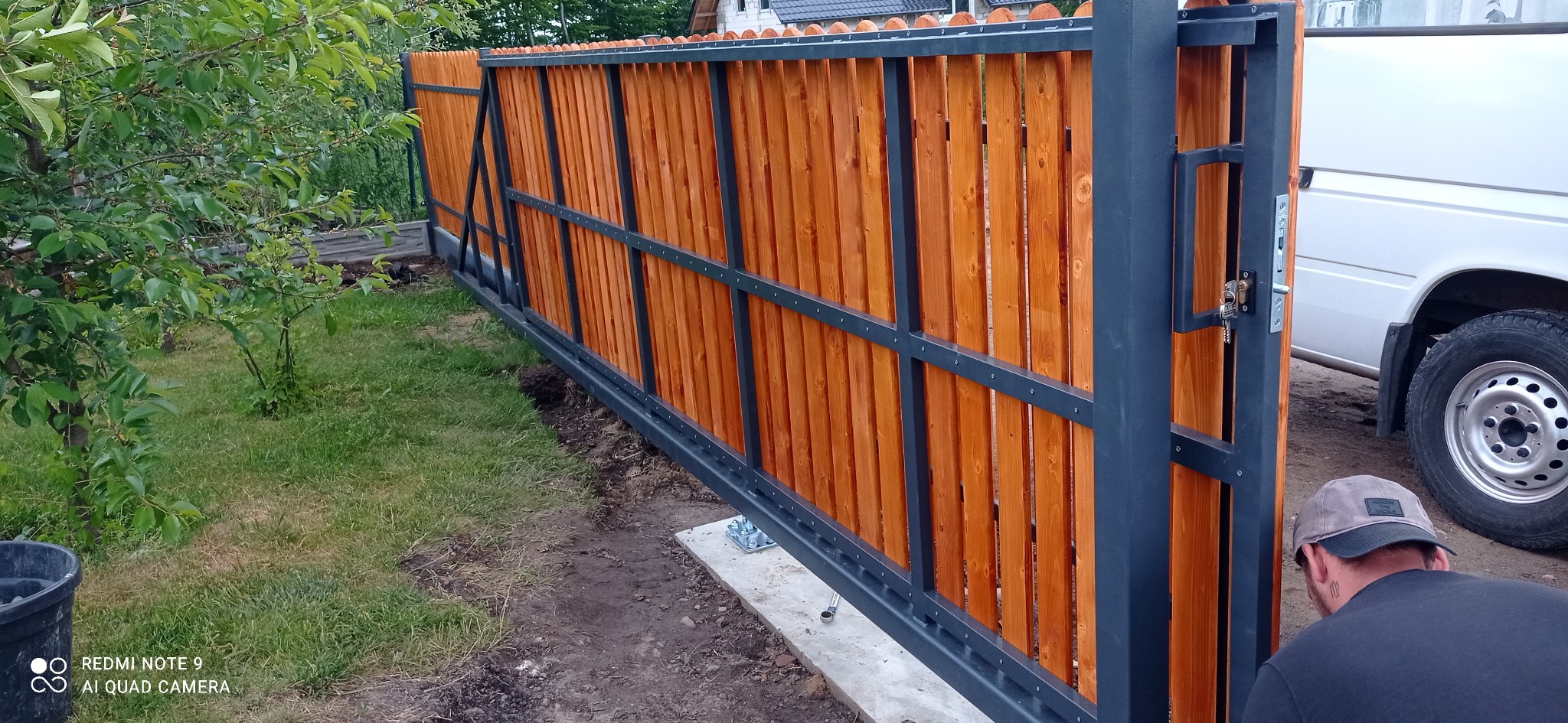 Przesuwna brama ogrodzeniowa z drewnianymi panelami w kolorze miodowym i ciemnoszarą metalową ramą, widoczna podczas montażu, z betonowym fundamentem i fragmentem białego samochodu w tle.