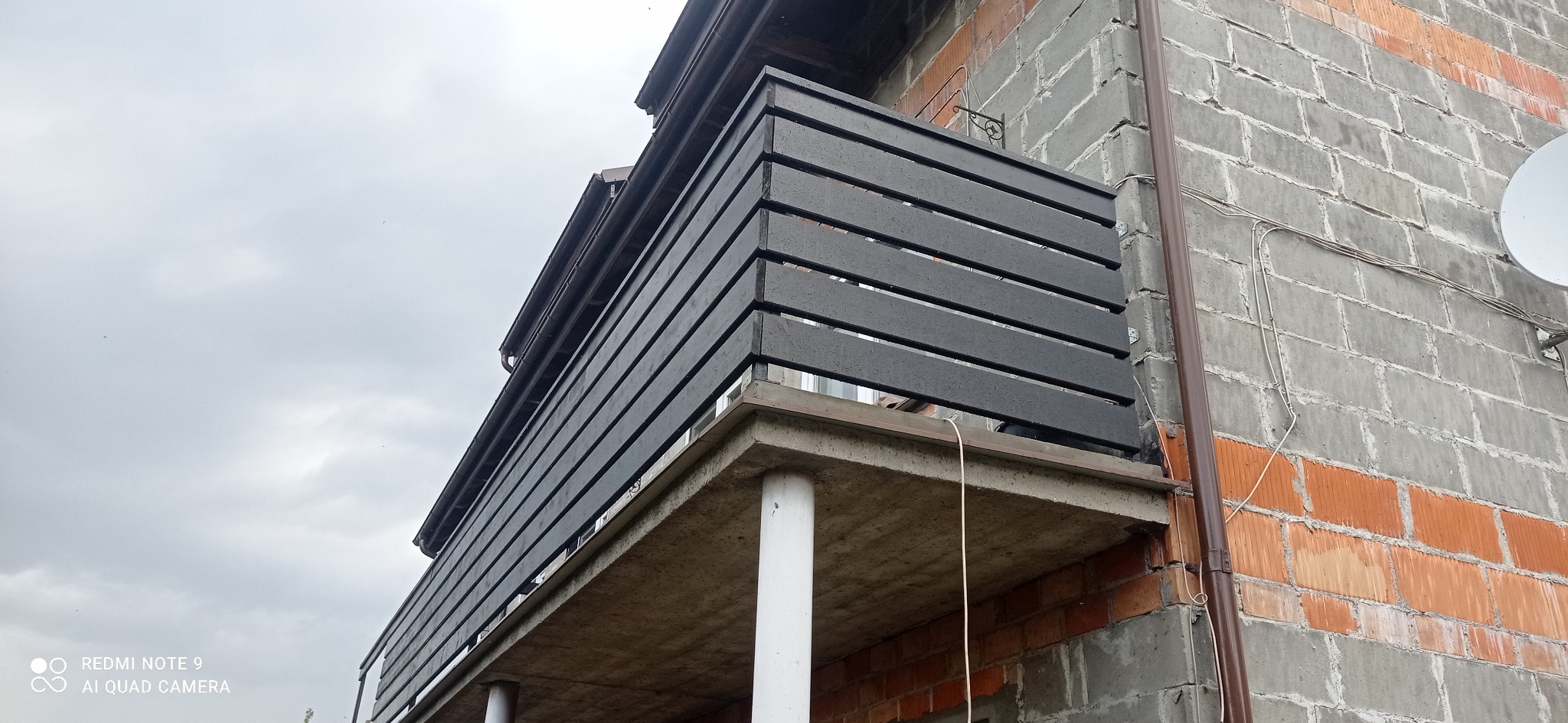 Czarny, drewniany balkon z poziomymi deskami, widoczny z dołu, na tle surowej ściany budynku z cegły i betonu.