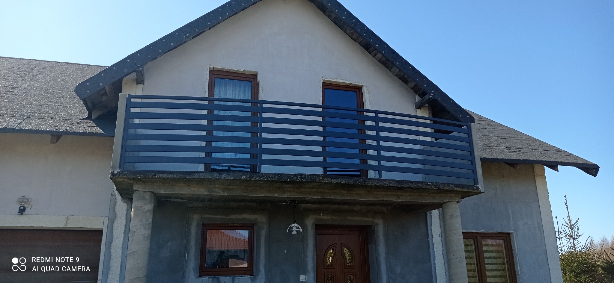 Szara, pozioma balustrada balkonowa z metalowych profili na tle elewacji domu w stanie surowym.