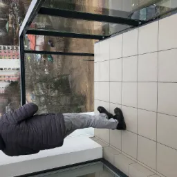 Glazura podłogowa balkonu z widokiem na osiedle miejskie przez szklaną balustradę, widoczne buty osoby stojącej na balkonie.