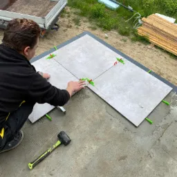 Układanie szarej płytki podłogowej na zewnątrz, z użyciem poziomicy, gumowego młotka i zielonych krzyżyków dystansowych, widok z góry na częściowo wykończoną powierzchnię.