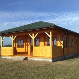 Żółty dom szkieletowy z zielonym dachem i brązowymi ramami okiennymi, widoczny z boku, z otwartą werandą i słupami w kształcie litery V, na tle pola pod błękitnym niebem.