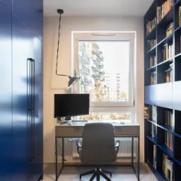 Nowoczesne biuro domowe z niebieską szafą, regałem na książki, szarym biurkiem i krzesłem, oświetlone lampą biurkową i światłem dziennym z okna z widokiem na budynek mieszkalny.