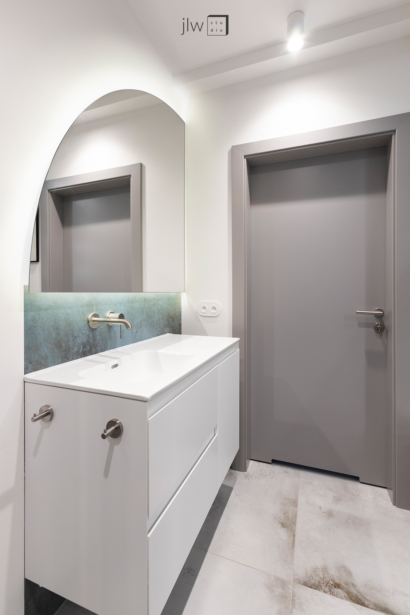 Nowoczesna łazienka z białą szafką umywalkową, okrągłym lustrem bez ramy, szarą ścianą z niebieskim akcentem nad umywalką i szarymi drzwiami po prawej stronie.