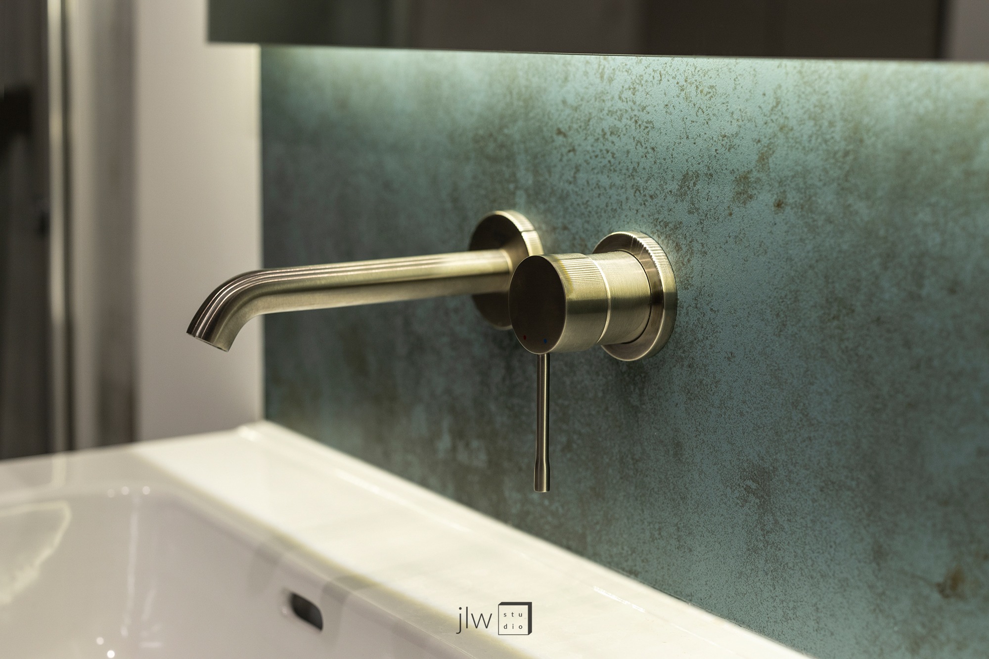 Bateria umywalkowa ścienna w kolorze szczotkowanego złota na tle ściany z efektem patyny, widoczna biała umywalka i logo JLW Studio