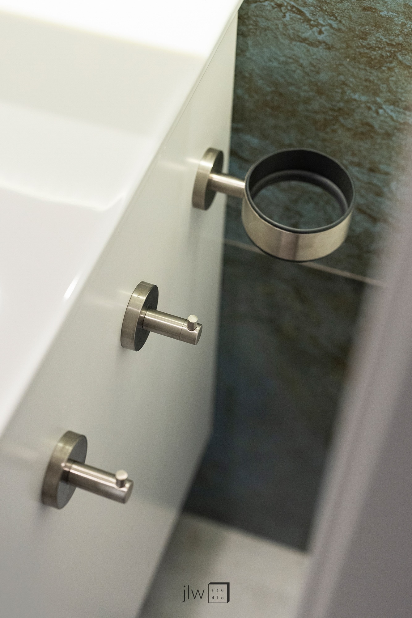 Biała szafka łazienkowa z trzema metalowymi uchwytami i uchwytem na kubek, na tle ściany z zielonego kamienia.