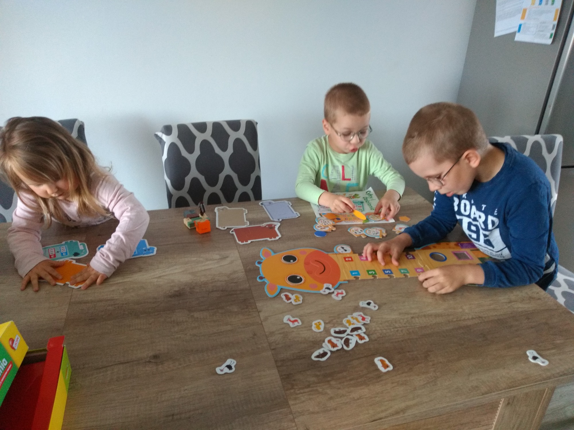Troje dzieci w okularach i bez, siedzi przy drewnianym stole układając puzzle i bawiąc się zabawkami edukacyjnymi, w tle szaro-białe krzesła i biała ściana.