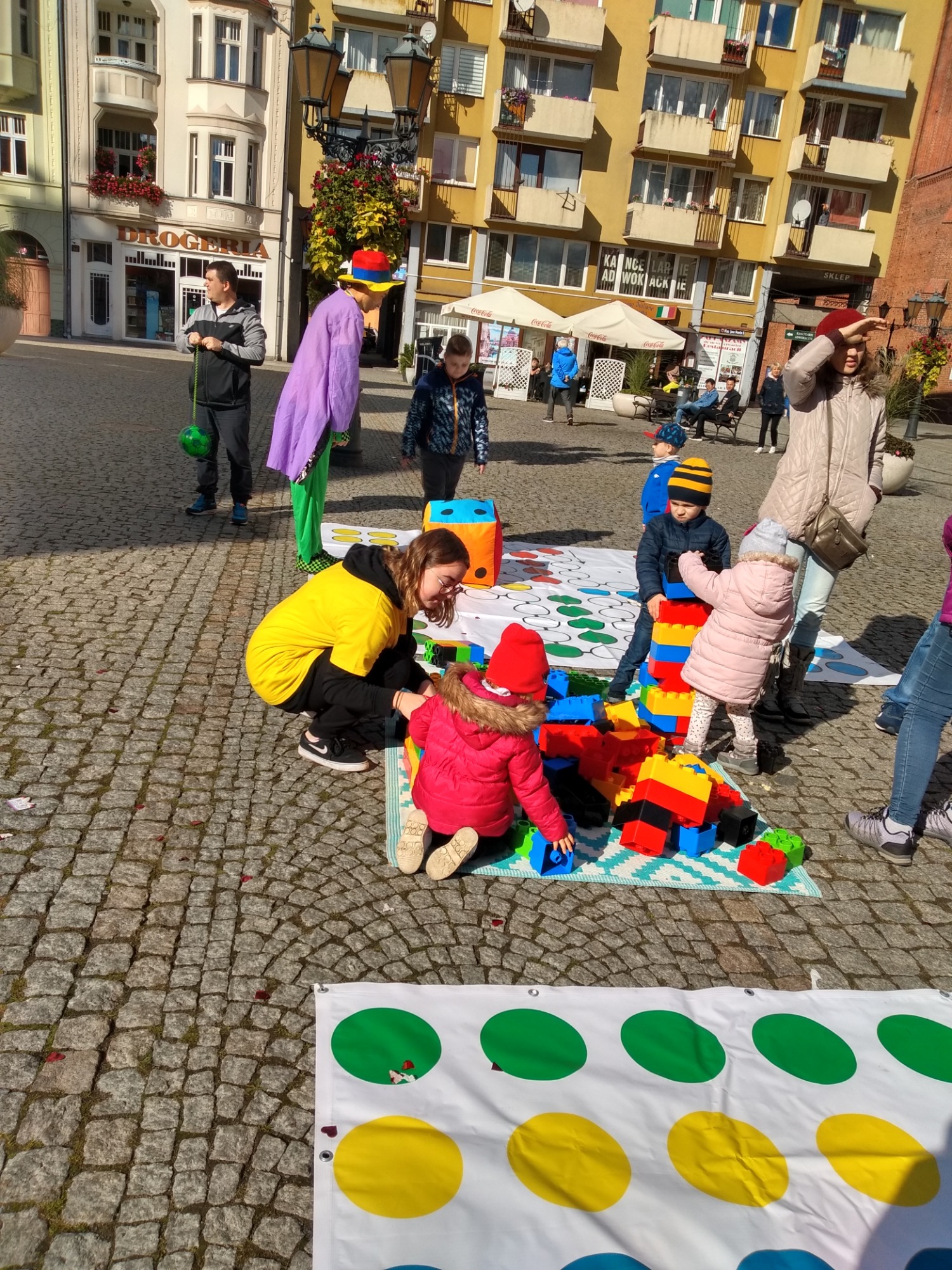 Animator w fioletowym płaszczu i kolorowym kapeluszu prowadzi gry i zabawy z dziećmi na placu miejskim, wykorzystując kolorowe klocki i matę do gry w Twister.