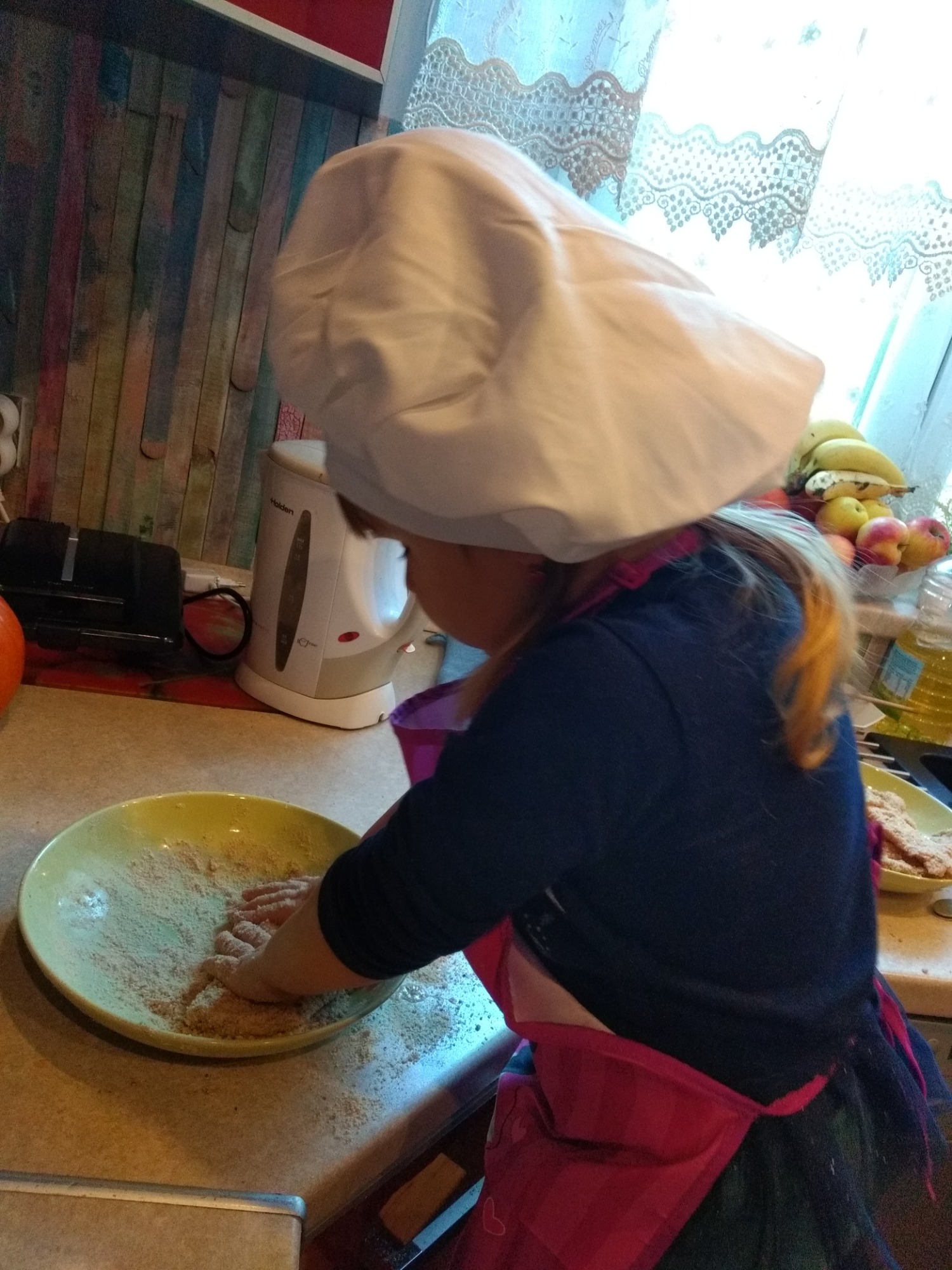 Dziecko w czapce kucharskiej obtacza kotlety w bułce tartej na żółtym talerzu w domowej kuchni z drewnianą ścianą i owocami w tle.