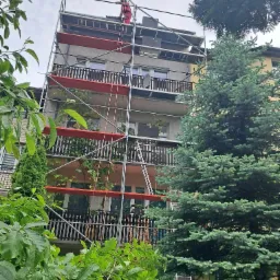 Pracownik w czerwonym kombinezonie na rusztowaniu przy remontowanym bloku z balkonami, widok z dołu przez zieleń drzew.