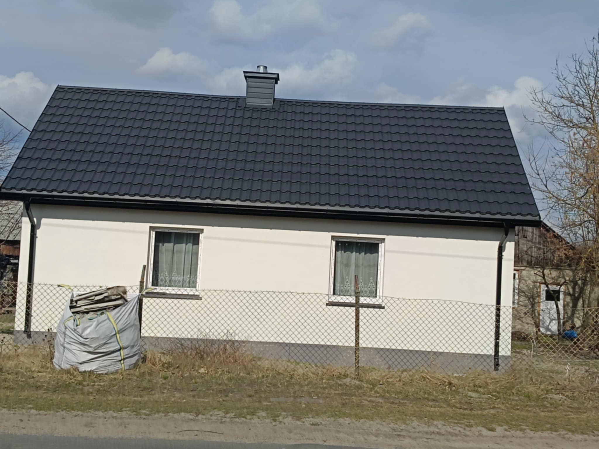 Dom jednorodzinny z nowym, ciemnoszarym dachem z blachodachówki i kominem, widoczny worek z odpadami budowlanymi przy ogrodzeniu.