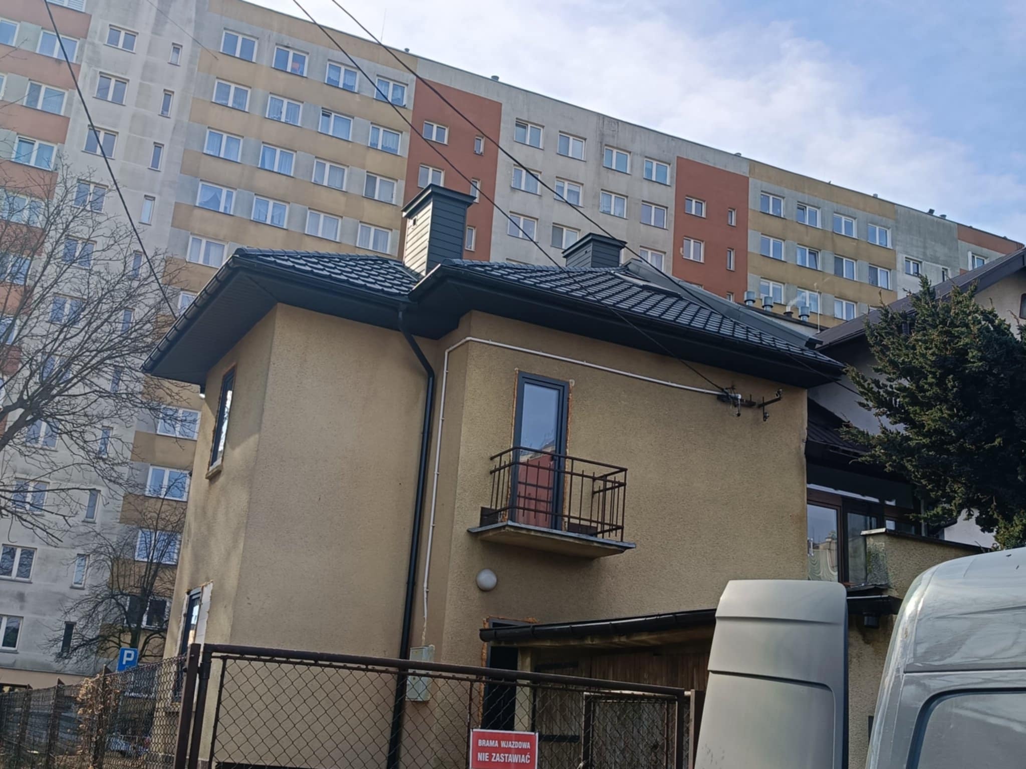 Dom jednorodzinny z ciemną dachówką na tle bloku mieszkalnego, widoczne kominy i fragment rynny.