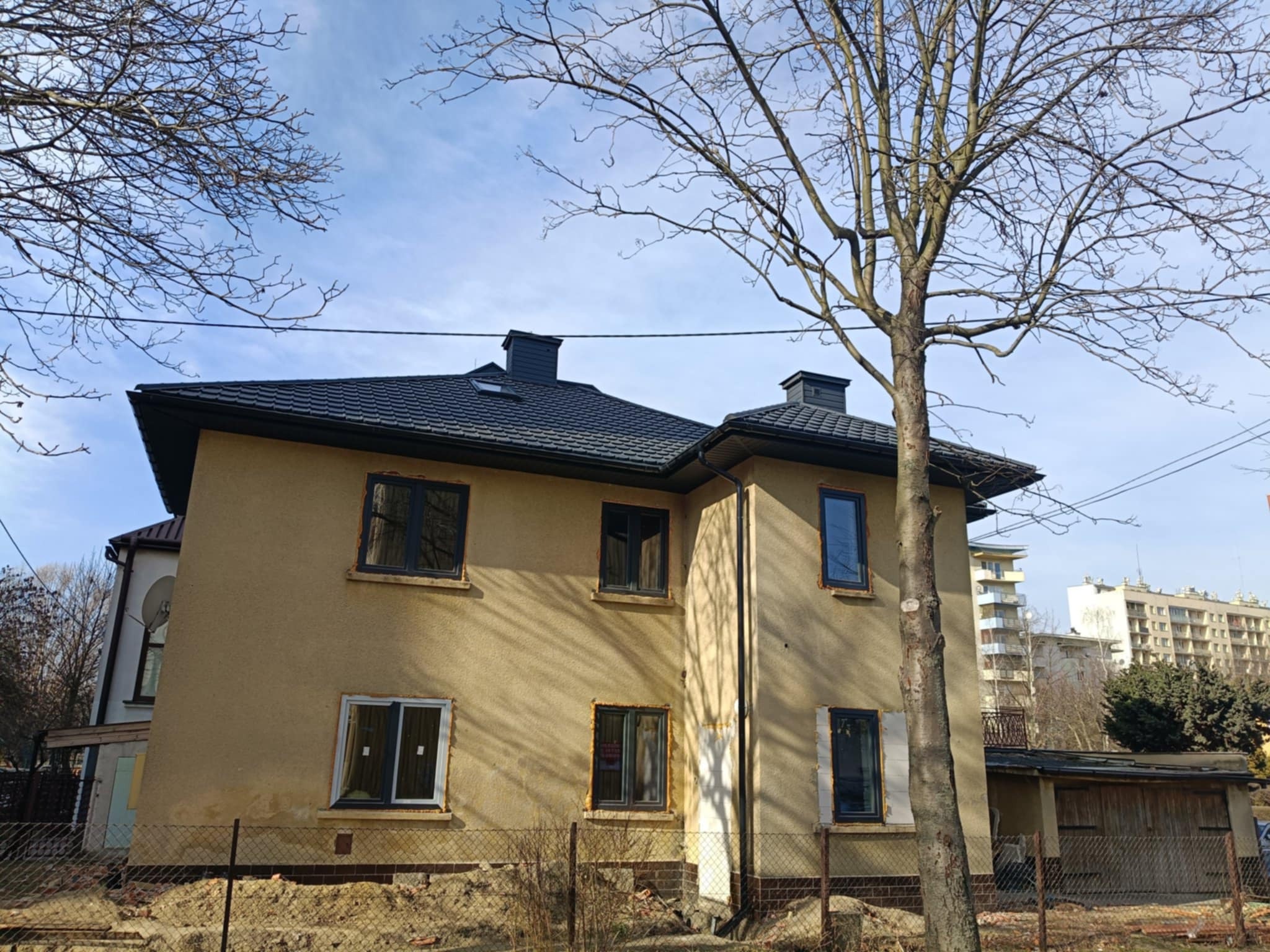 Dwupiętrowy dom z ciemnym dachem z dachówki i dwoma kominami, widoczny z poziomu gruntu za ogrodzeniem z siatki. Elewacja w odcieniach beżu. W tle budynki mieszkalne.