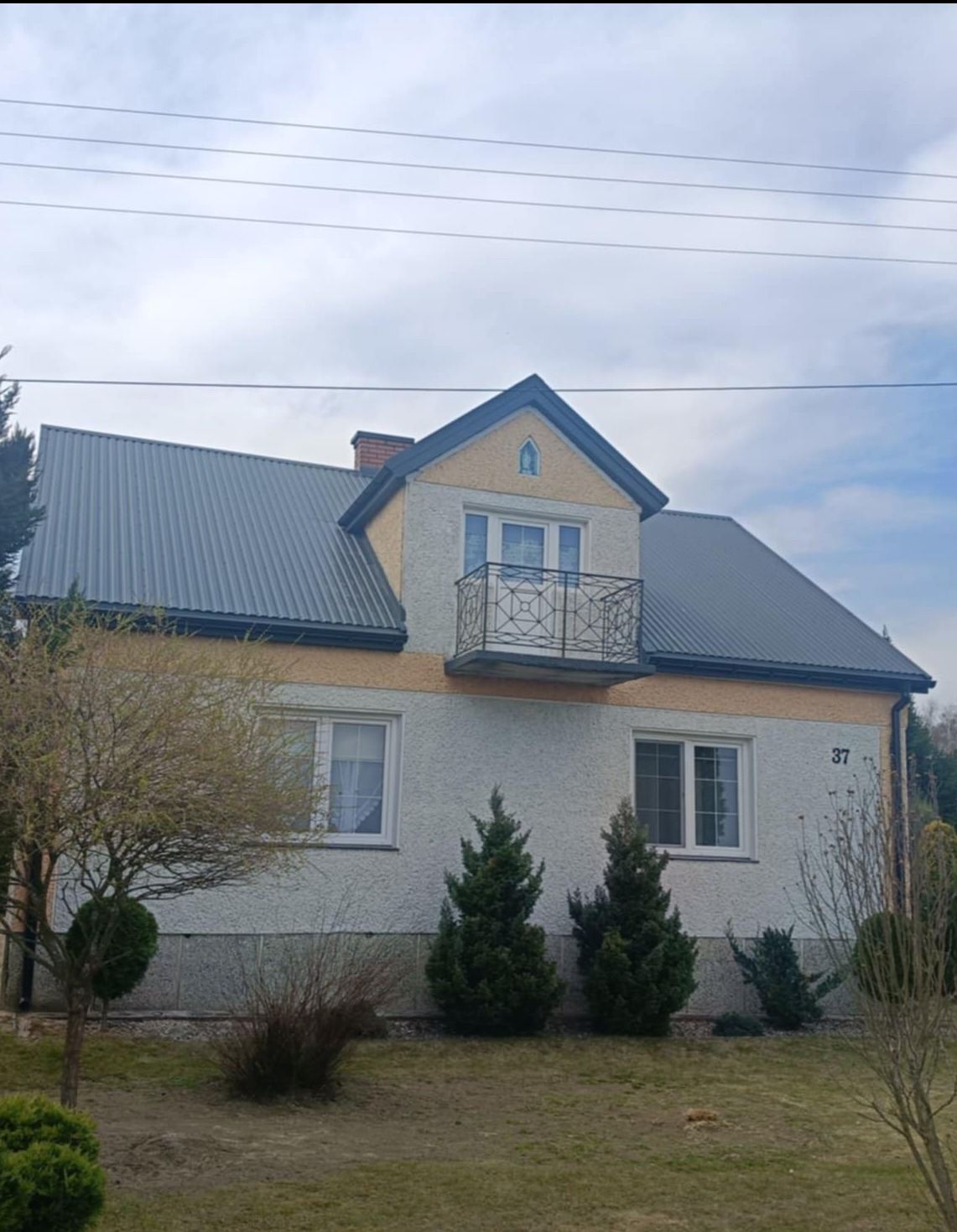 Dom jednorodzinny z nowym, ciemnoszarym dachem z blachy trapezowej, balkonem z czarną balustradą i numerem 37 na elewacji, otoczony zielenią.