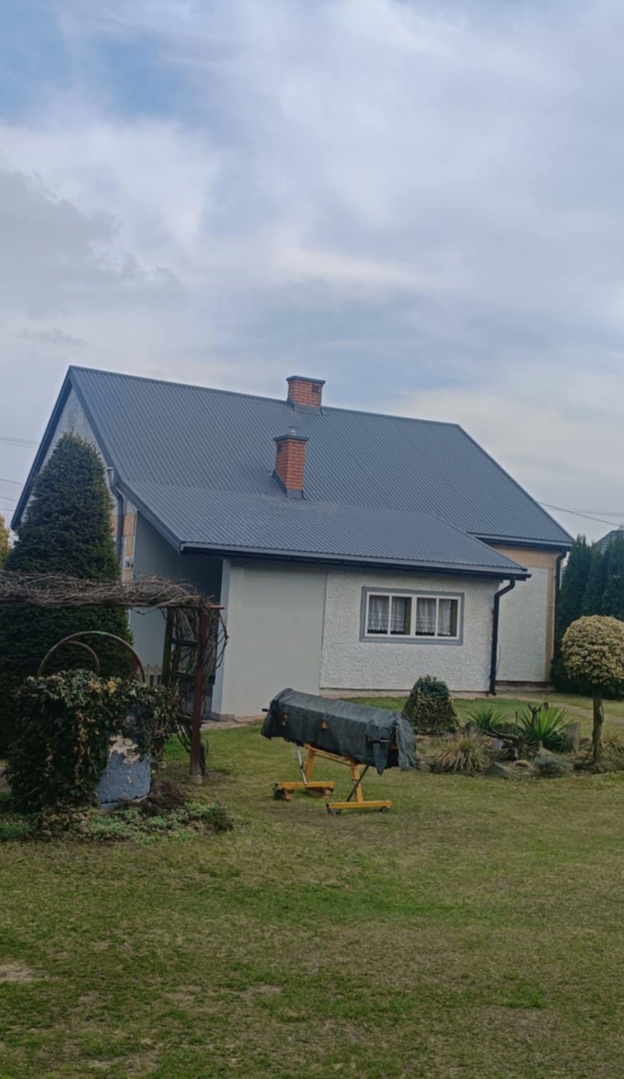 Dom jednorodzinny z nowym, ciemnoszarym dachem z blachy trapezowej, widoczne dwa kominy z cegły, trawnik z elementami dekoracyjnymi i osłoniętym rusztowaniem w tle.