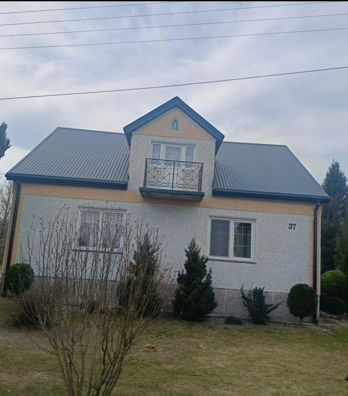 Dom jednorodzinny z nowym, szarym dachem z blachy trapezowej, widoczny balkon z kutą balustradą i numer domu 37.