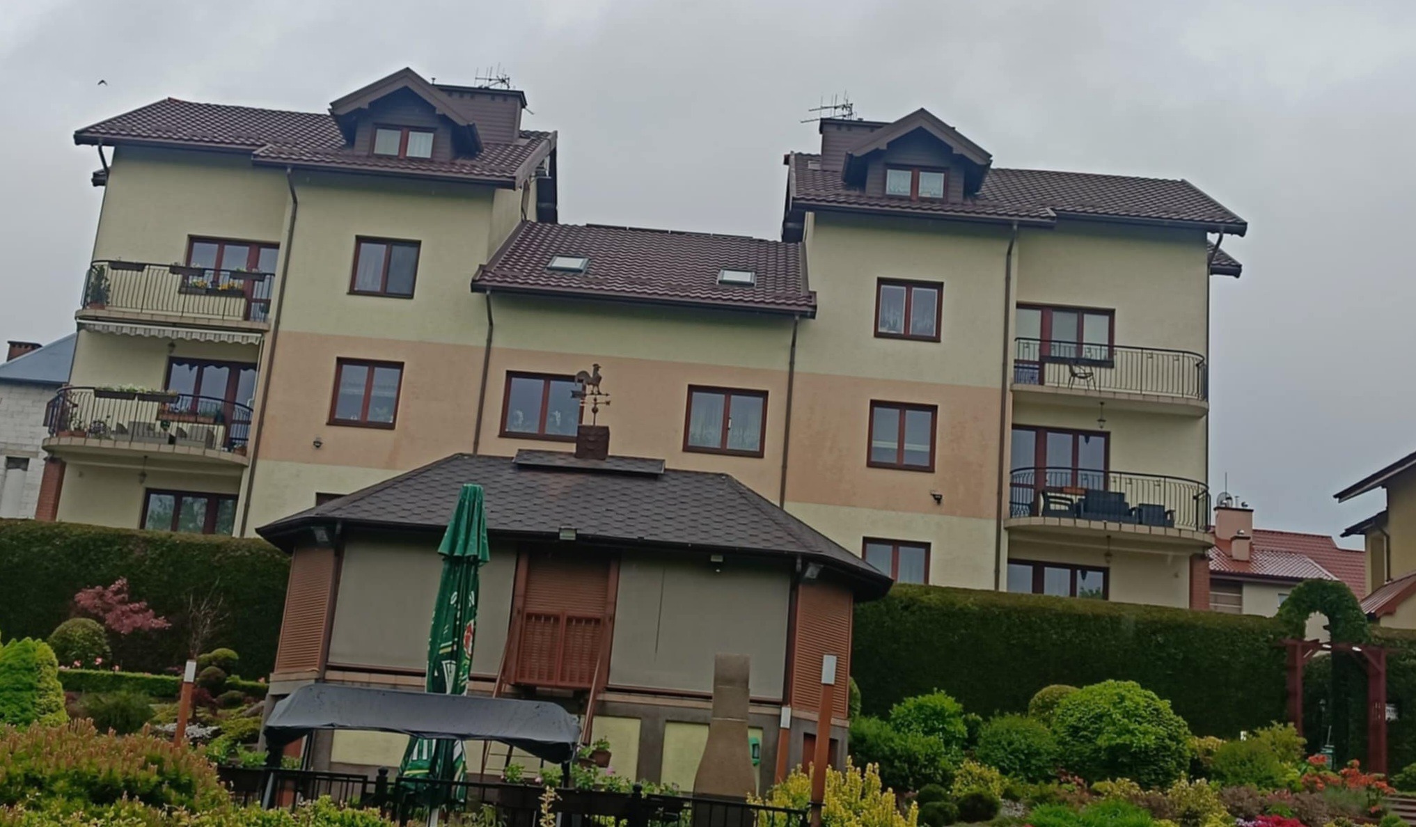 Dwa budynki wielorodzinne z brązowymi dachami i balkonami, widoczne zza altany ogrodowej z wiatrowskazem w kształcie koguta na dachu i zielonym parasolem.