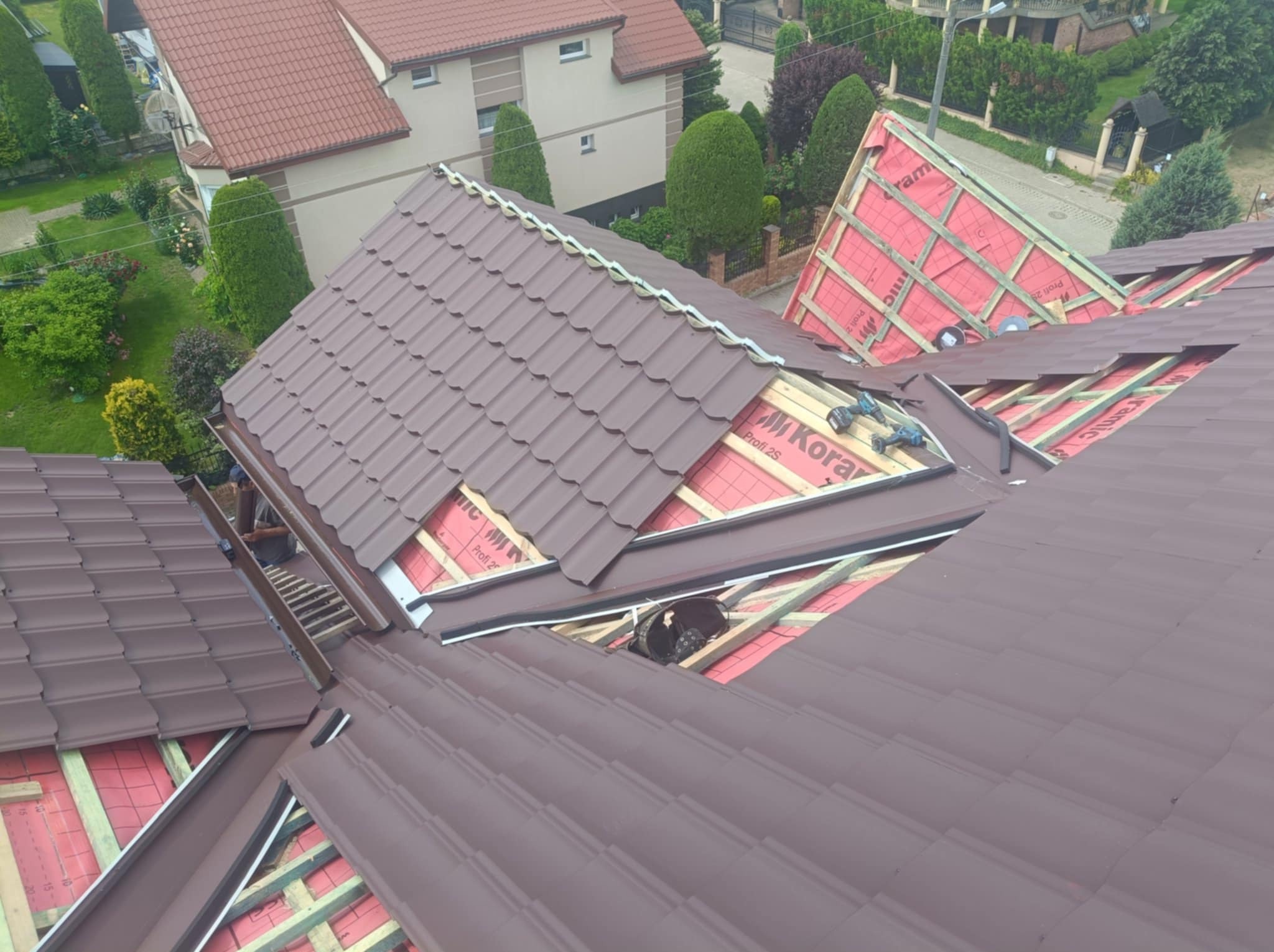 Układanie brązowej dachówki na połaci dachu z użyciem wkrętarki, widok z góry na dom w Zalesiu. Częściowo widoczna membrana dachowa i drewniana konstrukcja.