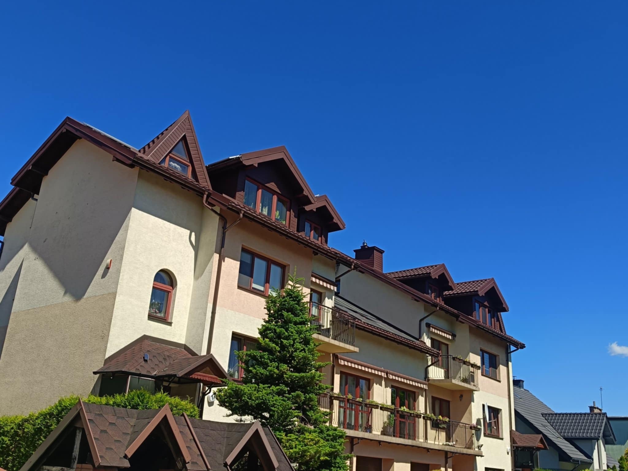 Elewacja budynku wielorodzinnego z brązowym dachem i rynnami, widziana z dołu pod kątem, w tle błękitne niebo. Widoczne balkony z kwiatami.