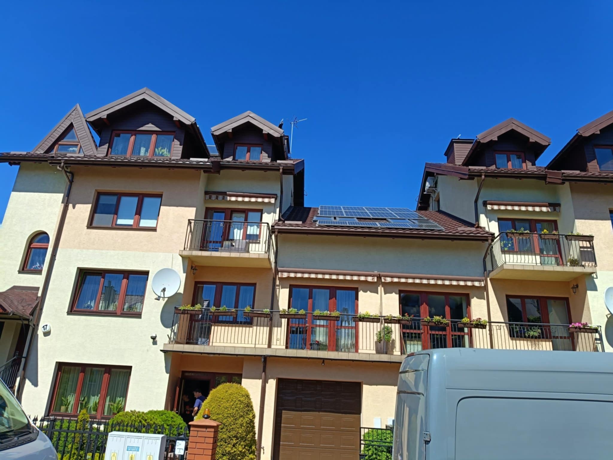 Trzy segmenty domu z brązowym dachem, panelami słonecznymi i balkonami. Błękitne niebo w tle, częściowo zasłonięte przez samochód dostawczy.