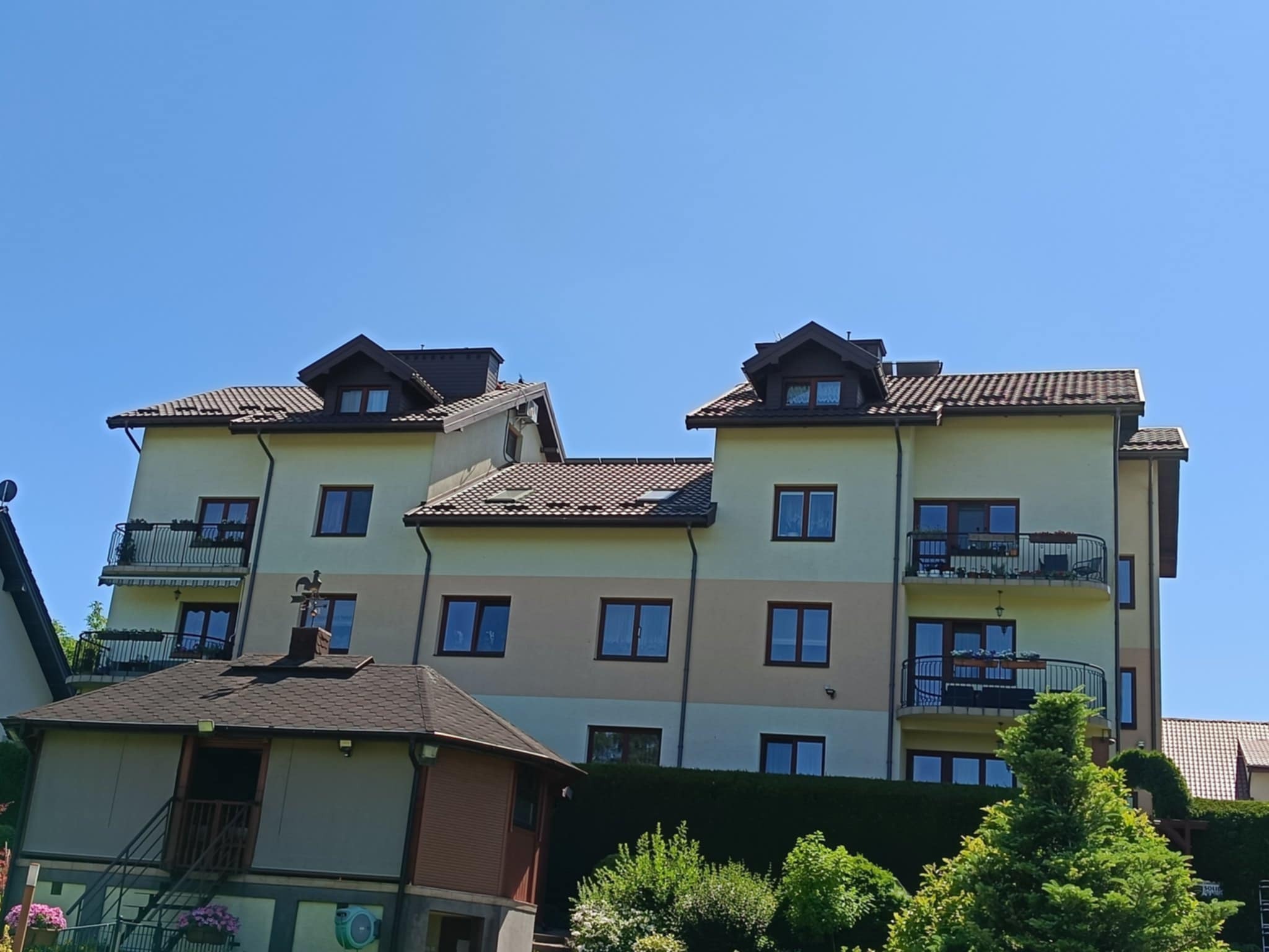 Widok na budynek wielorodzinny z brązowym dachem i balkonami, otoczony zielenią. Na pierwszym planie mały budynek z ciemnym dachem. Błękitne niebo w tle.