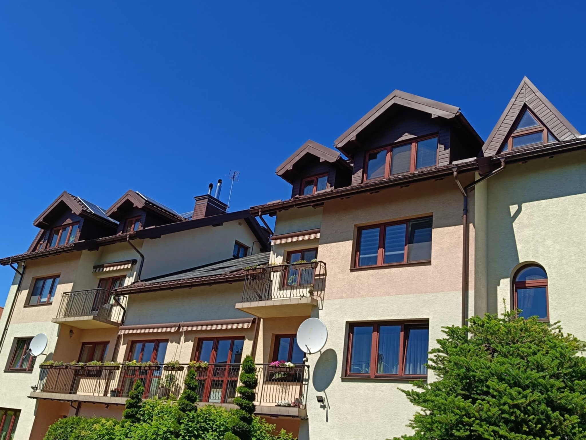 Wielopiętrowy budynek z balkonami i brązowym dachem na tle błękitnego nieba. Widoczne okna z brązowymi ramami i rośliny na balkonach.